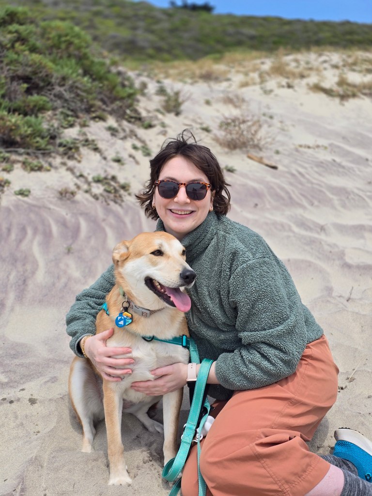 A woman in a green fleece sweater and orange pants kneels on a sandy beach, wearing dark sunglasses, holding a golden-brown, medium sized dog.
